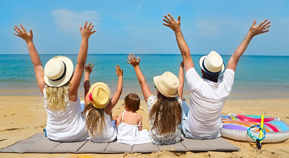 Fünfköpfige Familie mit Sonnenhüten auf dem Kopf, sitzen am Strand und strecken die Hände in die Luft 