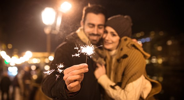 Junges Paar umarmt sich und feiert Silvester. Der Mann hält eine Sprühkerze in der Hand.