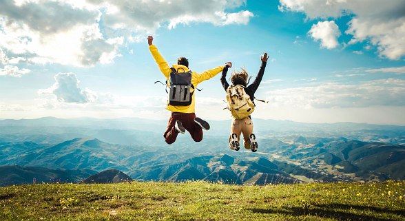 Ein Mann und eine Frau springen mit ihren Wanderrucksäcken in die Höhe und schmeißen die Hände in die Luft