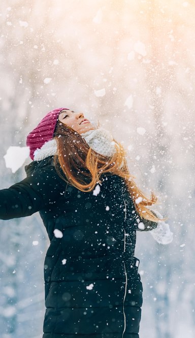 Frau mit warmer Jacke, Schal, Mütze und Handschuhen schaut lächelnd nach oben und es schneit. Im Hintergrund verschneite Bäumen auf Piste. Im Hintergrund schneebedeckte Bäume