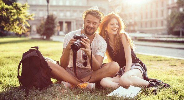 Junge Frau und junger Mann sitzen auf der Wiese in einer Stadt und sehen sich Fotos auf der Camera an