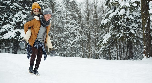 Mann trägt Frau auf dem Rücken und geht durch eine Winterlandschaft mit Wald im Hintergrund 