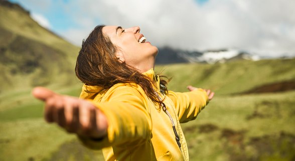 Junge Frau mit gelber Jacke, breitet die Arme aus und lächelt. Im Hintergrund Hügel und blauer Himmel mit Wolken