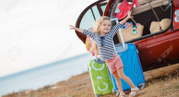 Kleines Mädchen streckt sich und lächelt. Im Hintergrund ein blauer und grüner Koffer und der offene Kofferraum eines Autos mit Urlaubsgepäck.