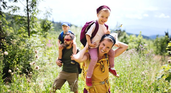 Familie beim Wandern. Eltern tragen ihre Kinder auf den Schultern und lächeln dabei.
