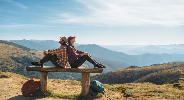 Paar sitzt auf einer Bank, Rücken an Rücken. Berge und Sonne im Hintergrund
