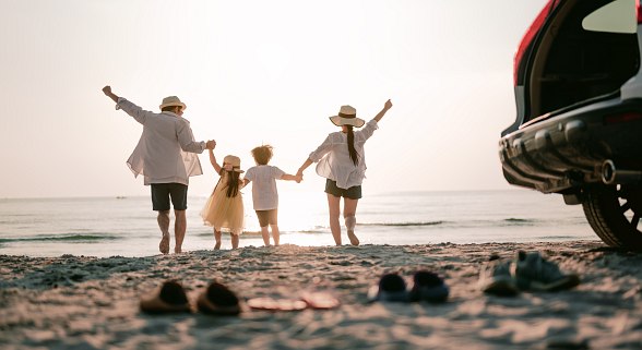 Familie am Strand mit Blick aufs Meer. Ein Mann, 2 Kinder und eine Frau halten sich an den Händen. Der Hinterteil eines Autos ist sichtbar. 