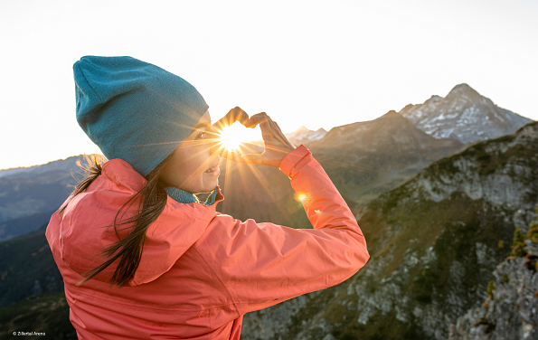 Frau zeigt Herz mit den Händen in die Sonne im Hintergrund mit Bergen